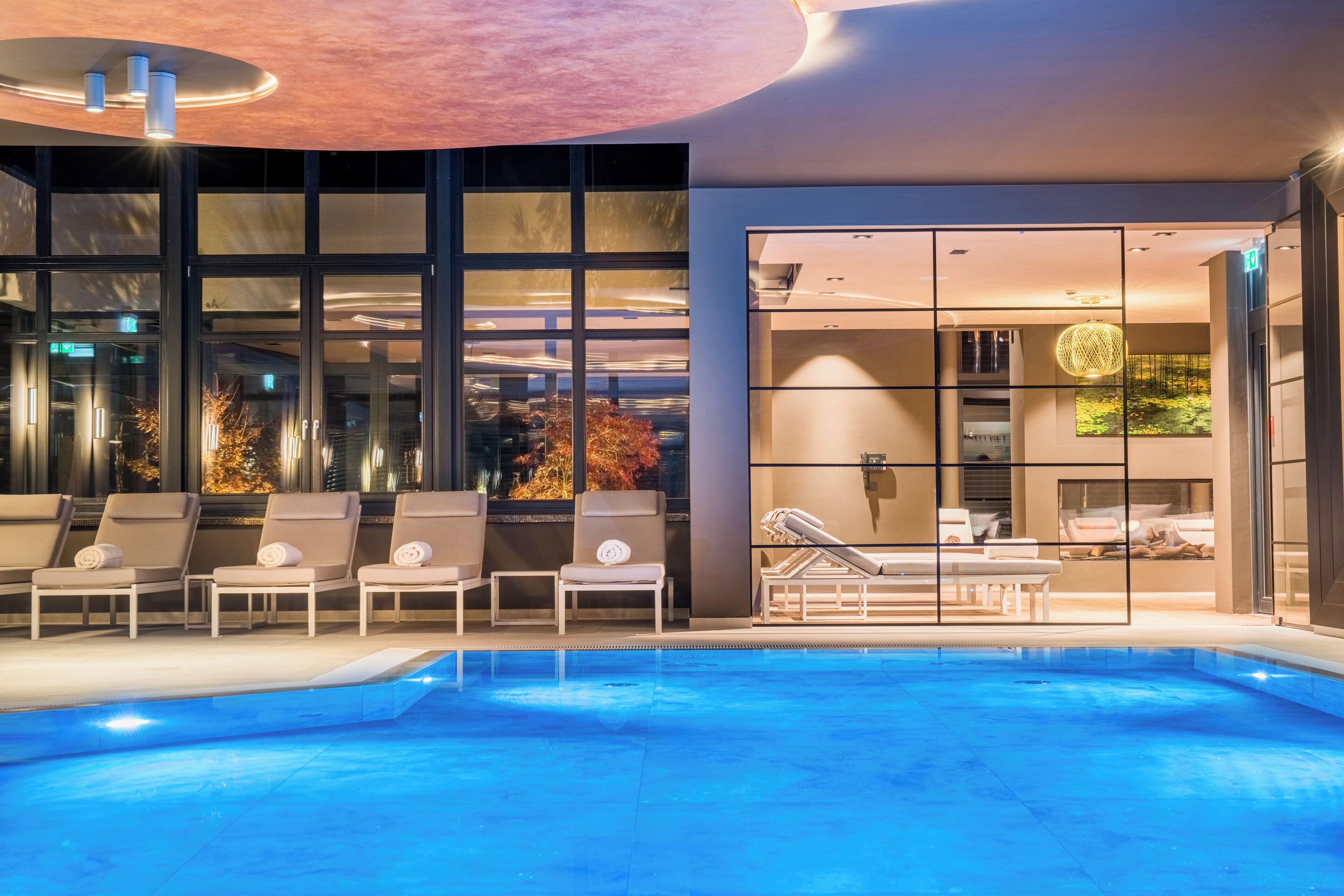 Un área de piscina interior moderna con sillas de descanso y una pared de vidrio que ofrece vistas del paisaje urbano.