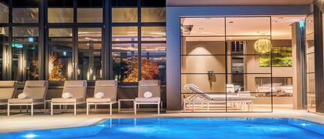 Un área de piscina interior moderna con sillas de descanso y una pared de vidrio que ofrece vistas del paisaje urbano.