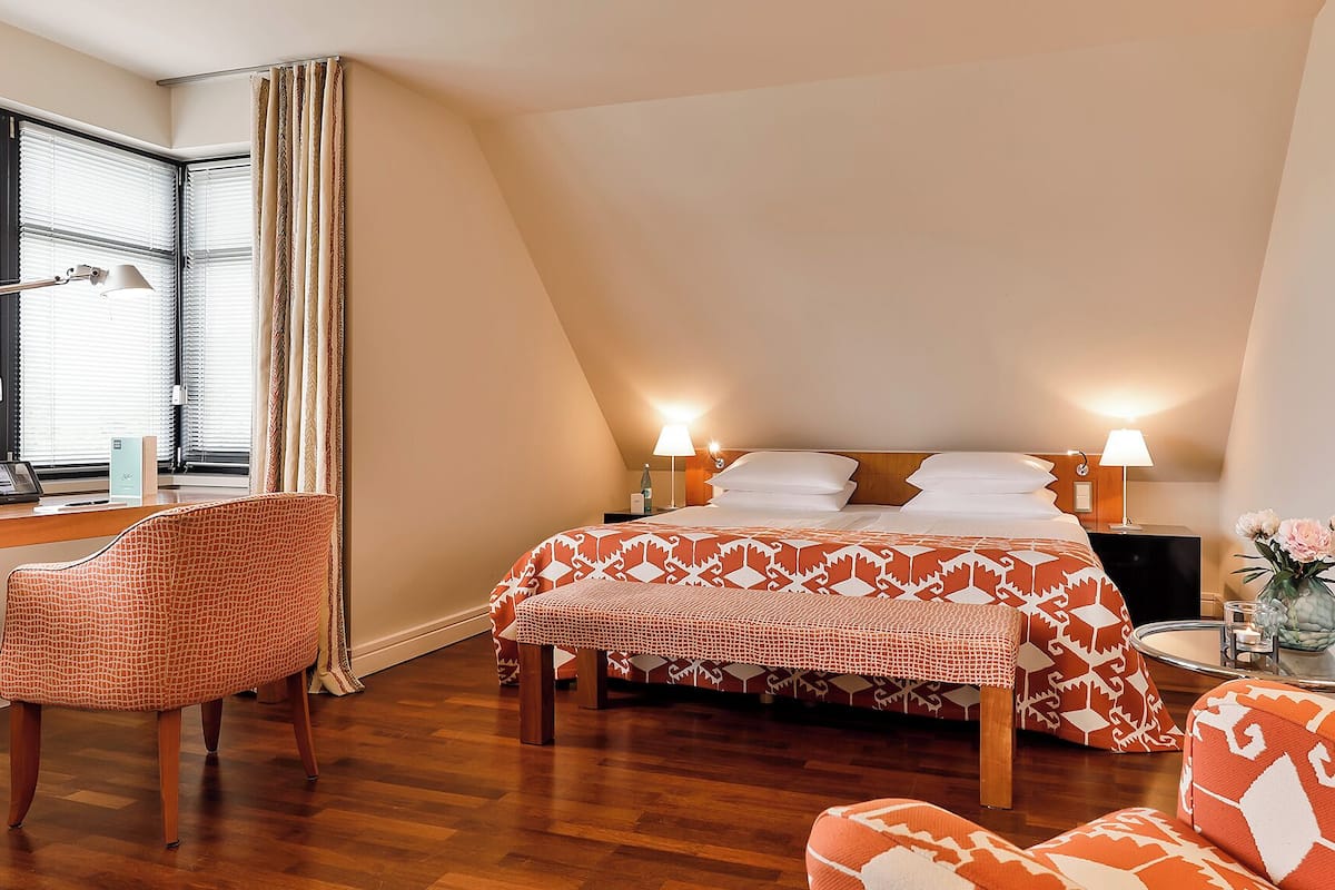 A hotel room with a bed, two chairs, a desk, and a window with blinds.