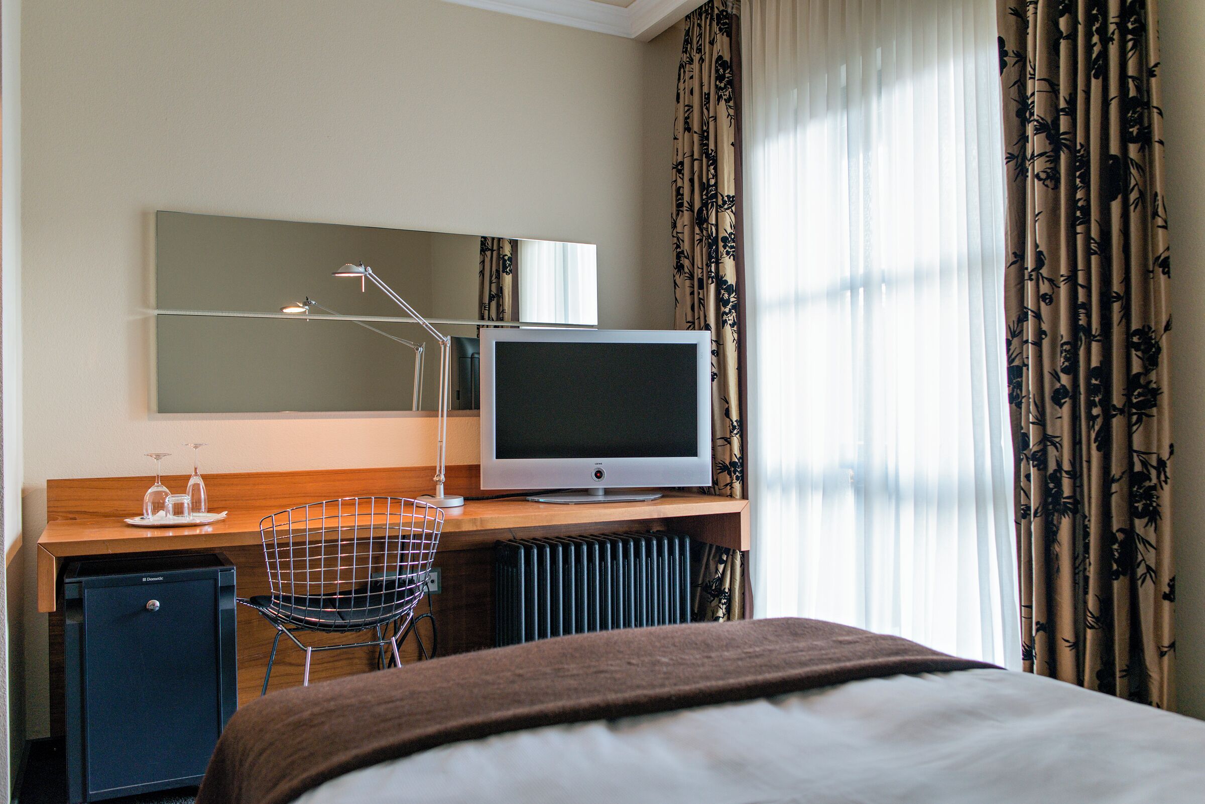 A hotel room with a bed, a desk with a television, a small refrigerator, a chair, and a large window with curtains.