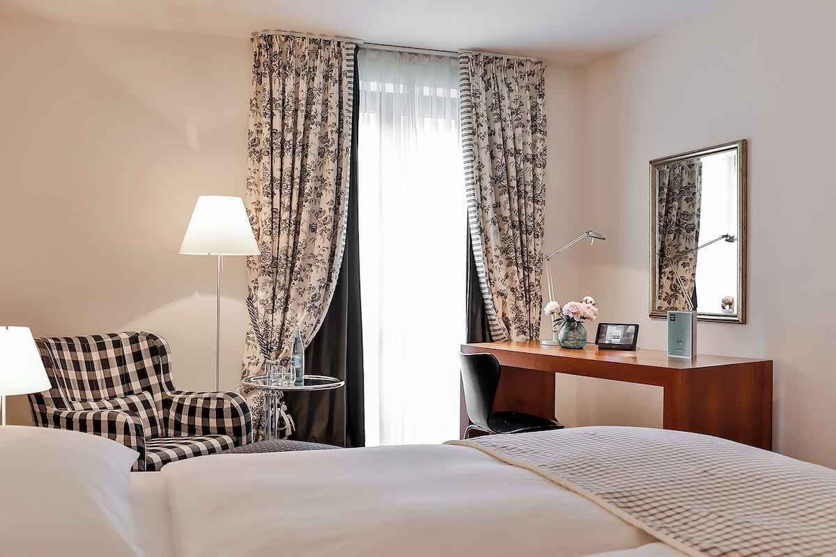 A hotel room with a bed, a checkered armchair, a desk with a lamp, a vase with flowers, and a large mirror.