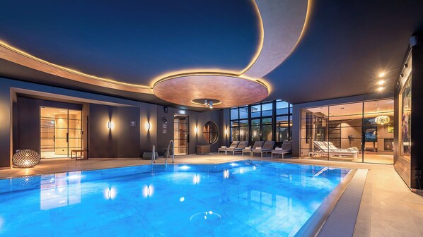 A modern indoor pool area with clear glass walls, lounge chairs, and a circular ceiling light fixture. - Zeller Hotel & Restaurant (Kahl)