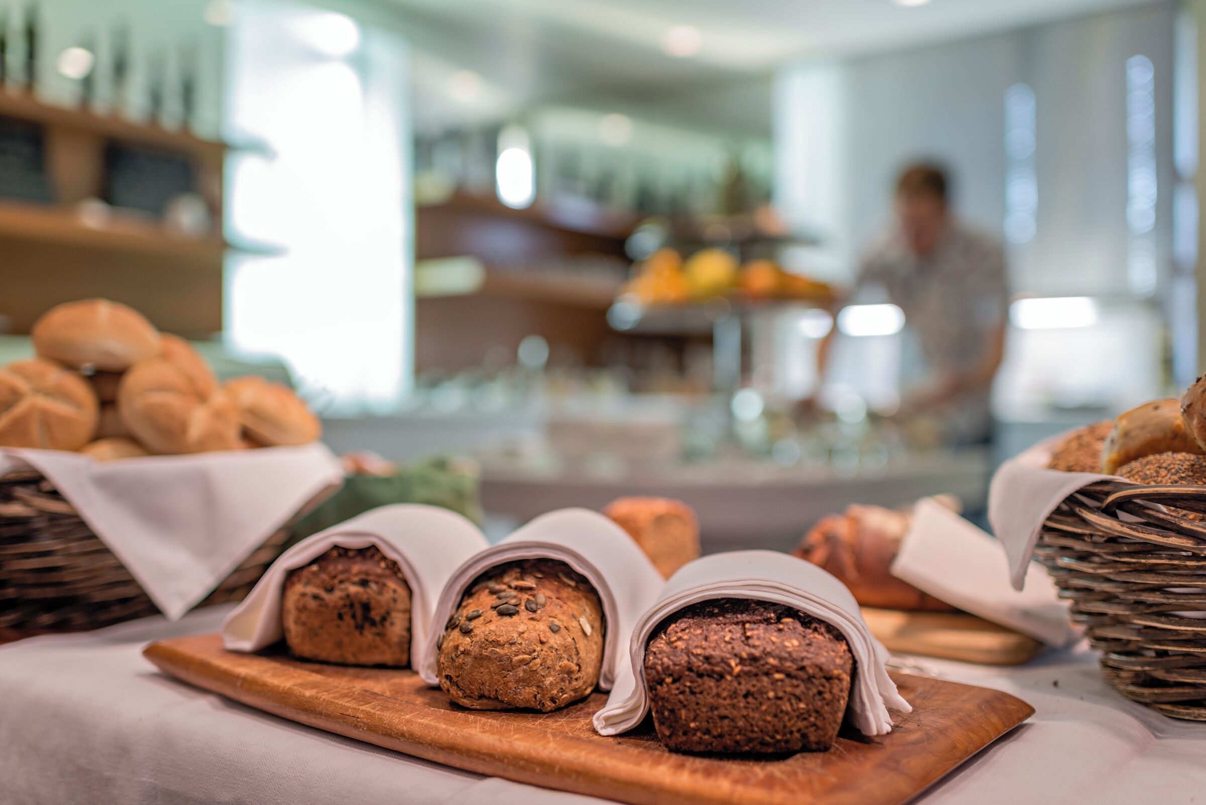A bakery display with breads and pastries on wooden boards, with a blurred background of a person and a counter.