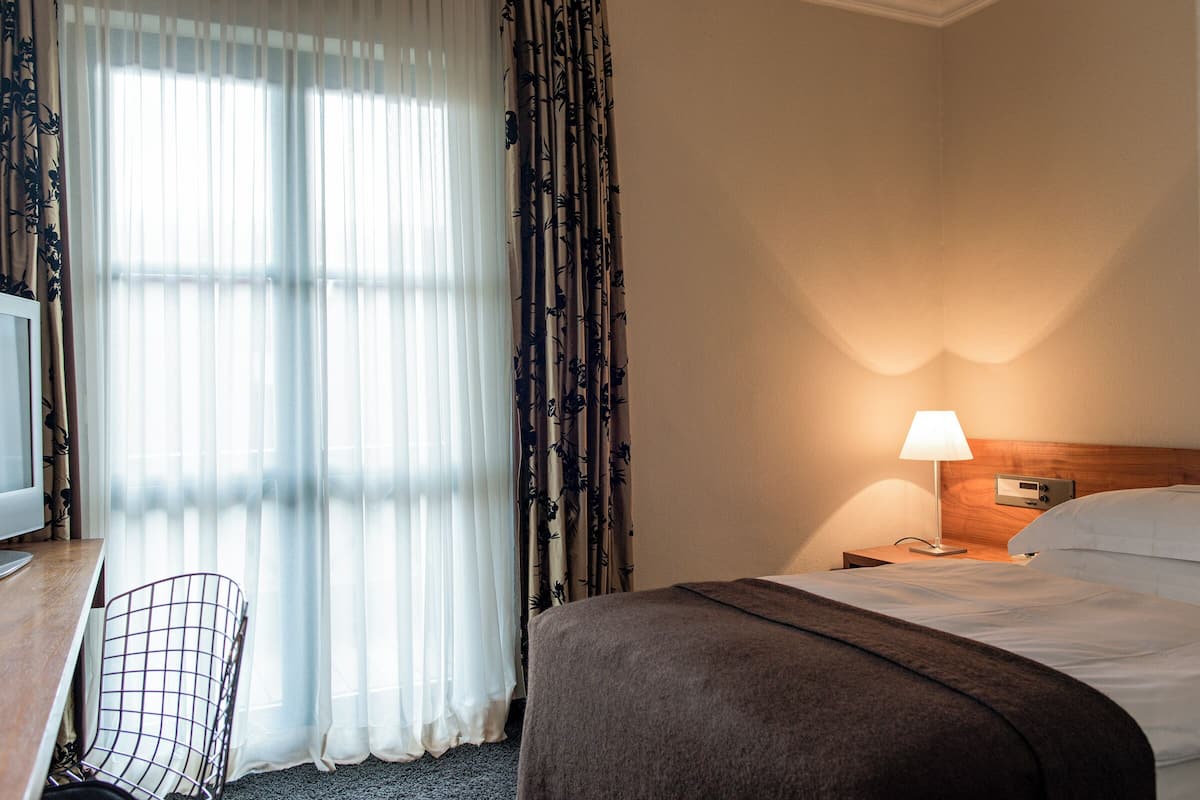A hotel room with a bed, a desk with a chair, a TV, and a nightstand with a lamp.