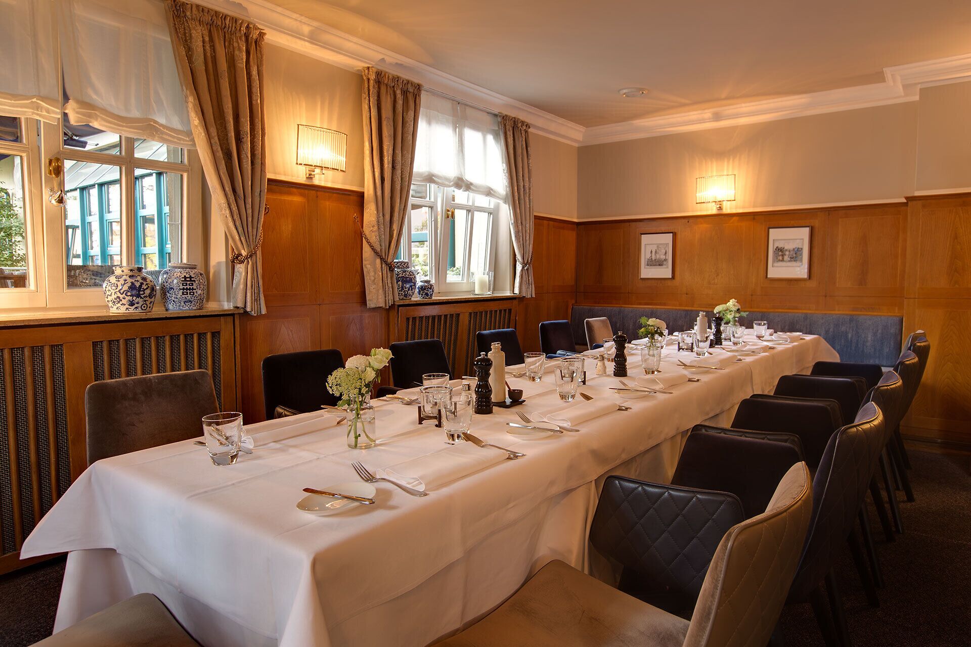 A long dining table set for a formal meal, with chairs and a view of the outdoors through large windows.