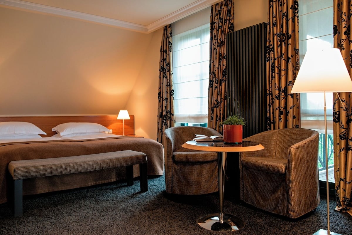 A hotel room with a bed, a bench, a chair, a small table with a plant, and a lamp.