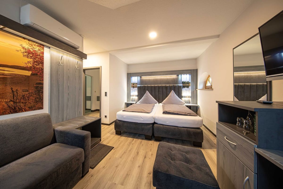 A modern hotel room with a bed, two bedside tables, a sofa, a TV, and a large window.