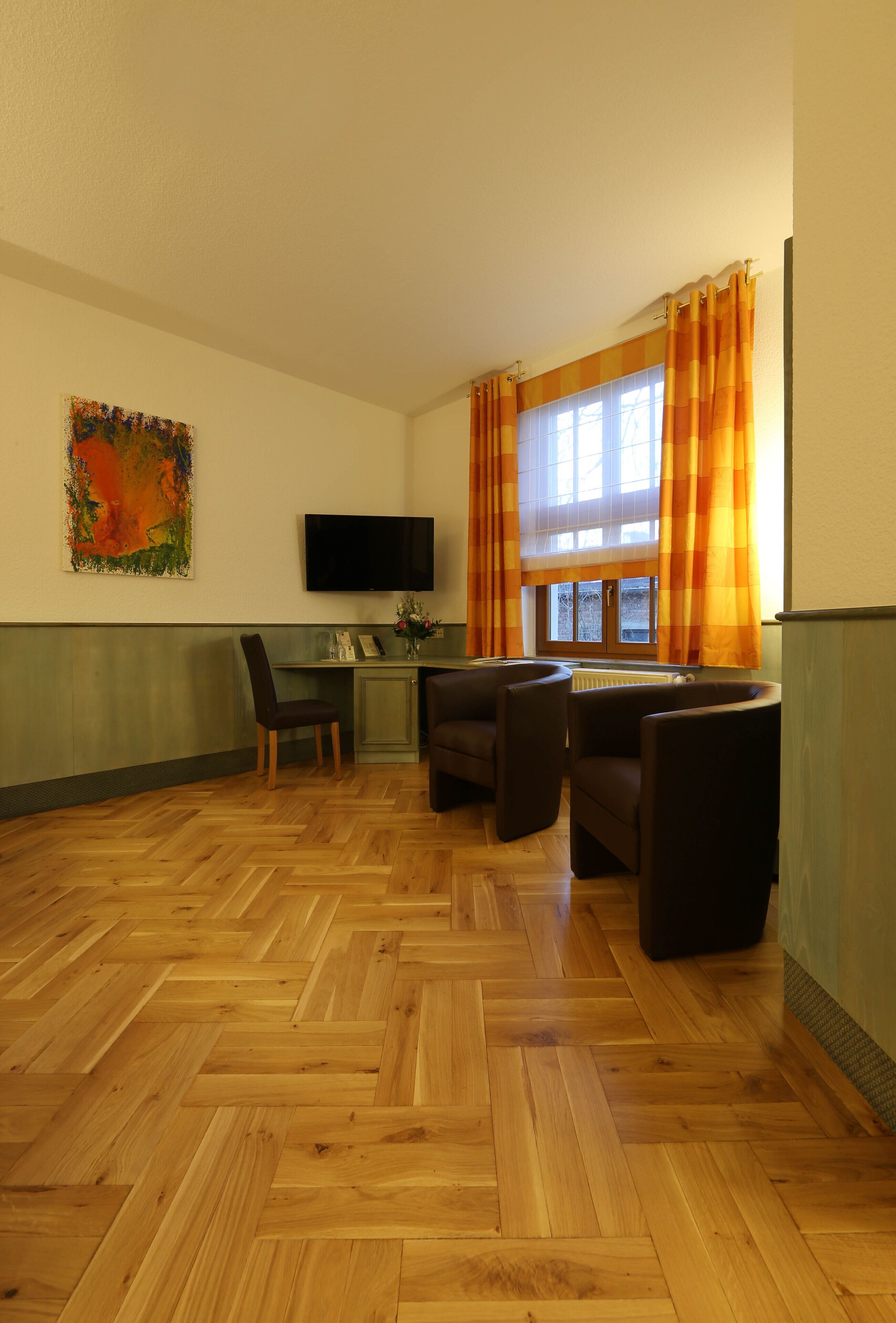 Una habitación con piso de madera en patrón de espiga, un televisor montado en la pared, un escritorio con una silla, y un sofá con sillones.
