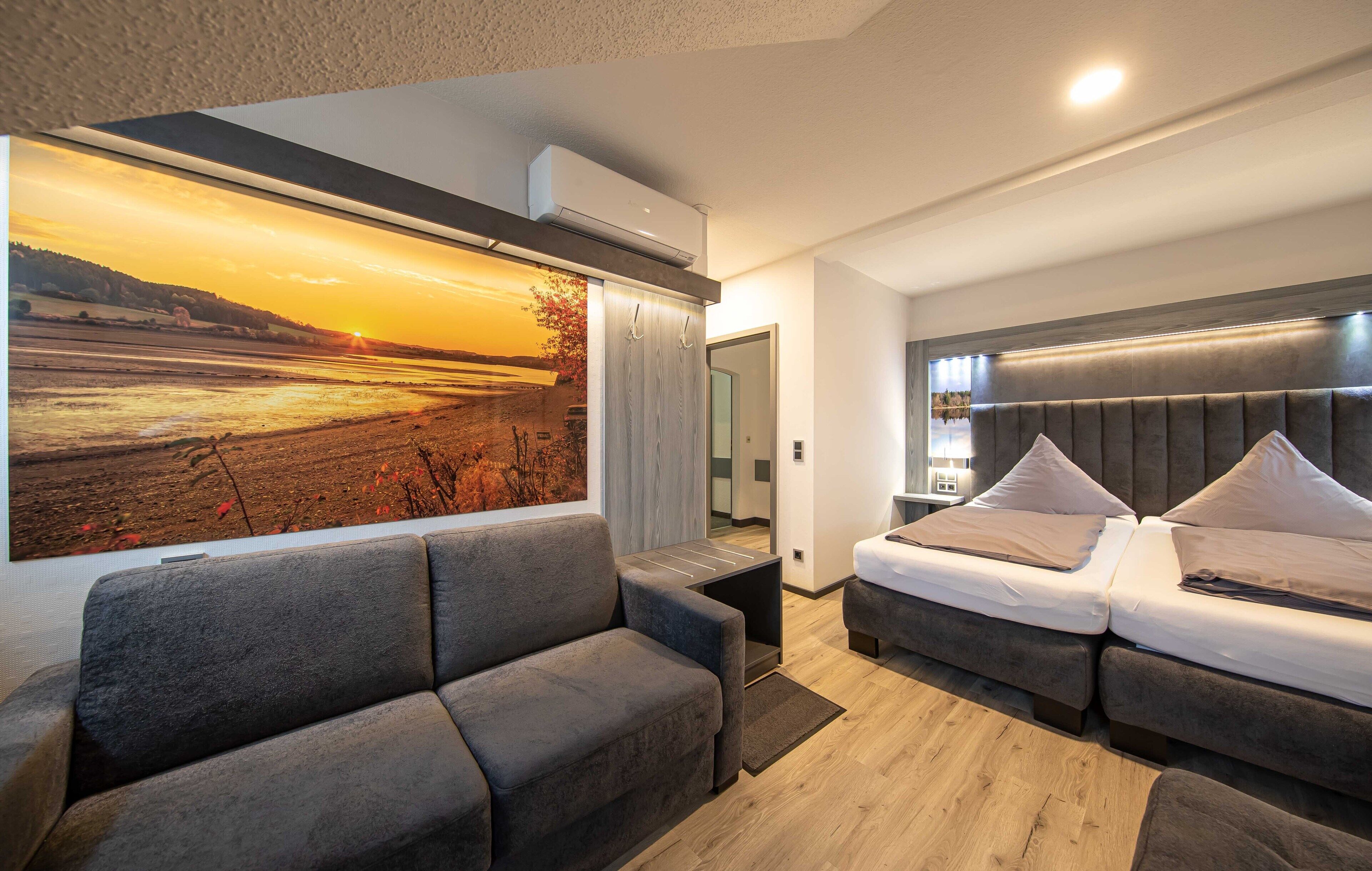A modern hotel room with a large painting of a sunset landscape, a sofa, a bed with a headboard, and a small table.