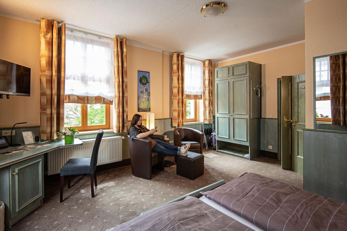 A hotel room with a desk, chair, bed, and a person using a laptop.