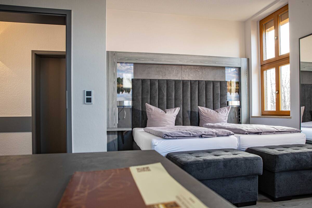 A modern bedroom with a large bed, two bedside tables, and a window with a view of a lake.