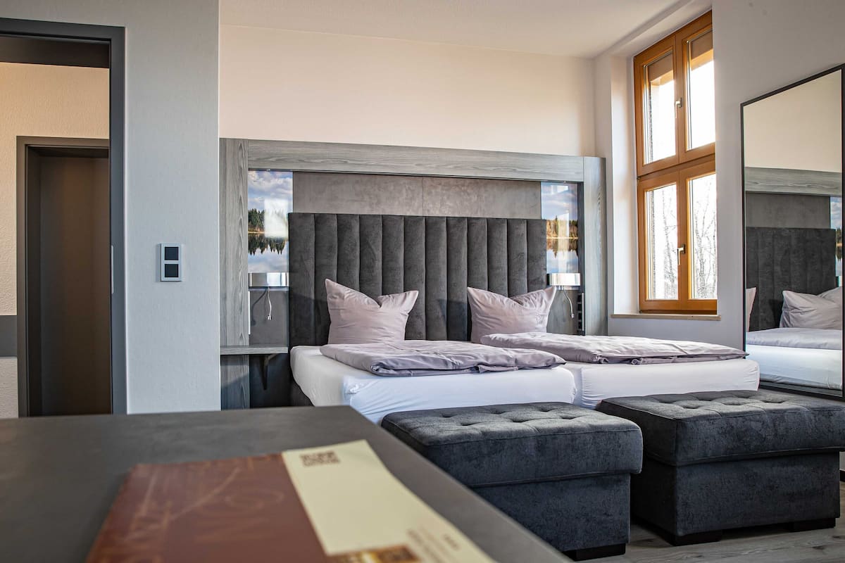 A modern hotel room with a large bed, a seating area with ottomans, a large window with a view, and a door with a light switch.