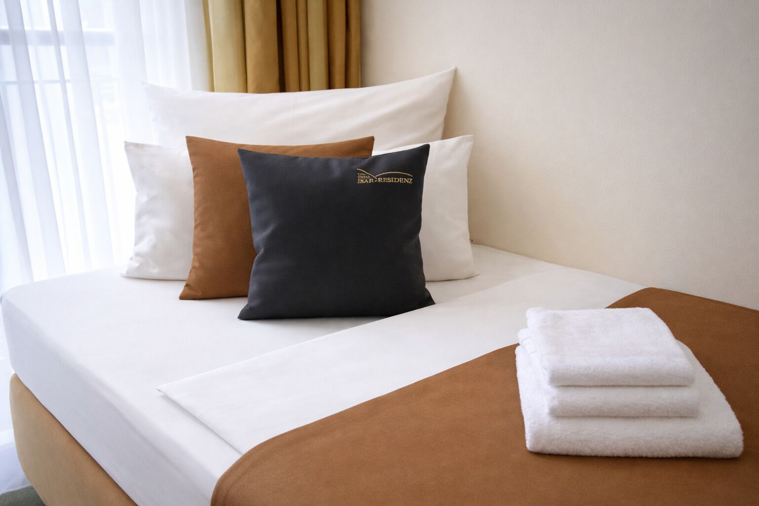 A neatly made bed with white sheets, a brown throw blanket, and a set of white towels.
