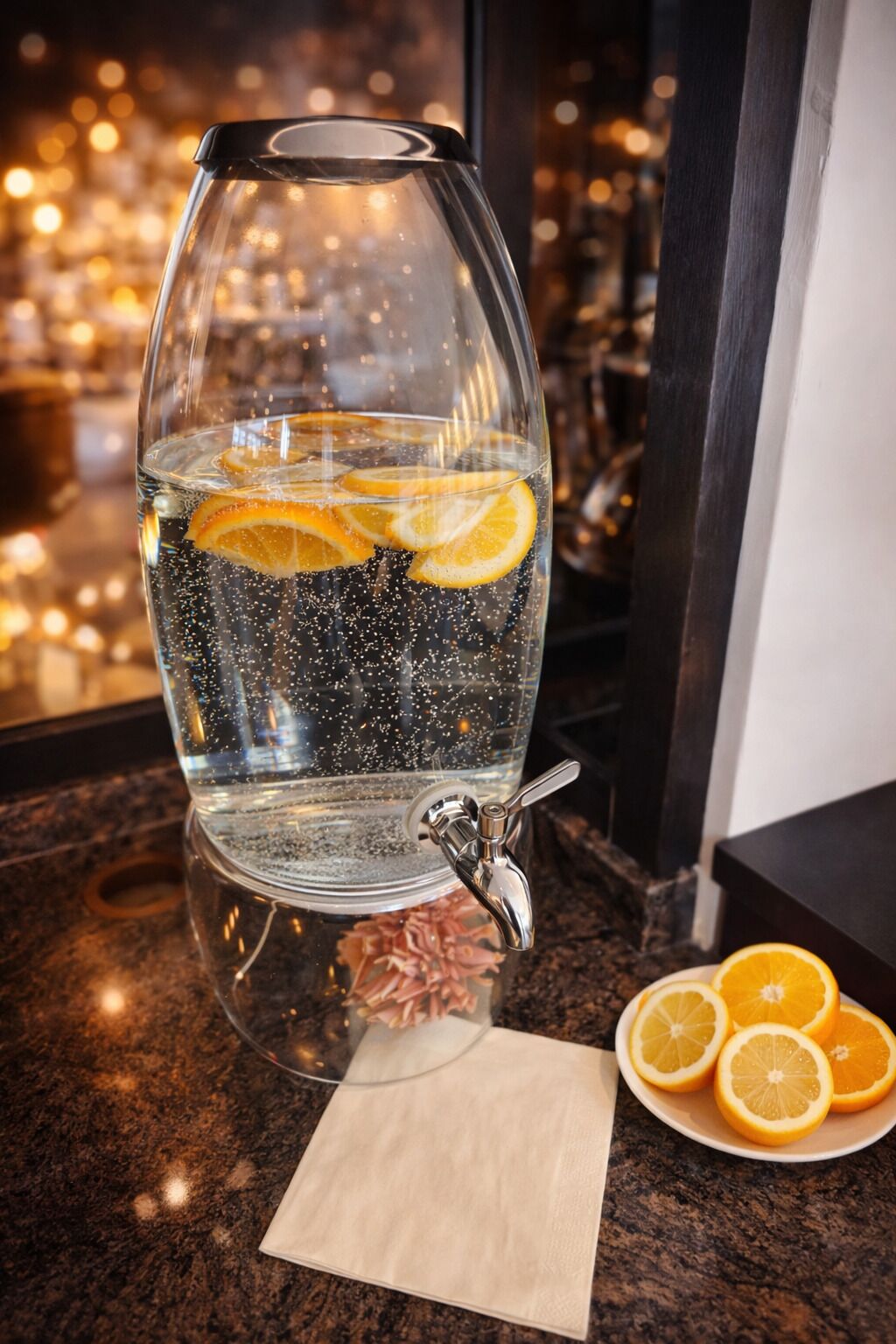 A glass pitcher with a faucet, filled with water and orange slices, on a granite countertop with a plate of orange slices beside it.