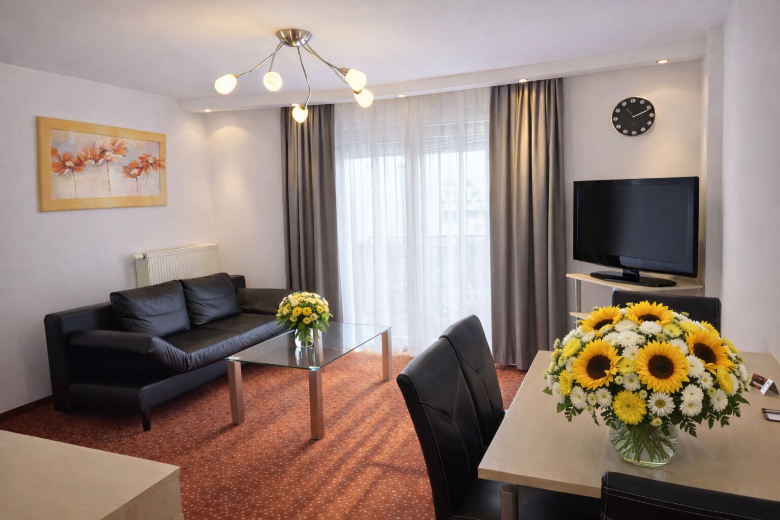 A modern hotel room with a black leather sofa, a glass coffee table, a dining table with a vase of sunflowers, a flat-screen TV, and a painting on the wall.