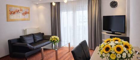 A modern hotel room with a black leather sofa, a glass coffee table, a dining table with a vase of sunflowers, a flat-screen TV, and a painting on the wall.