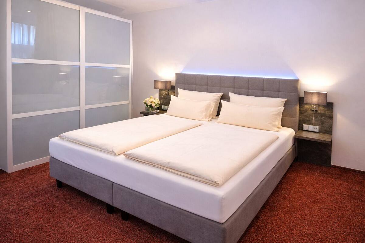 A double bed with a grey headboard, two bedside tables with lamps, a large window with frosted glass panels, and a red carpeted floor.