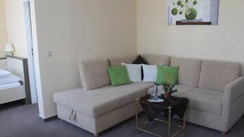 A living room with a beige sofa, green and white pillows, a small side table with a vase, and a painting on the wall.