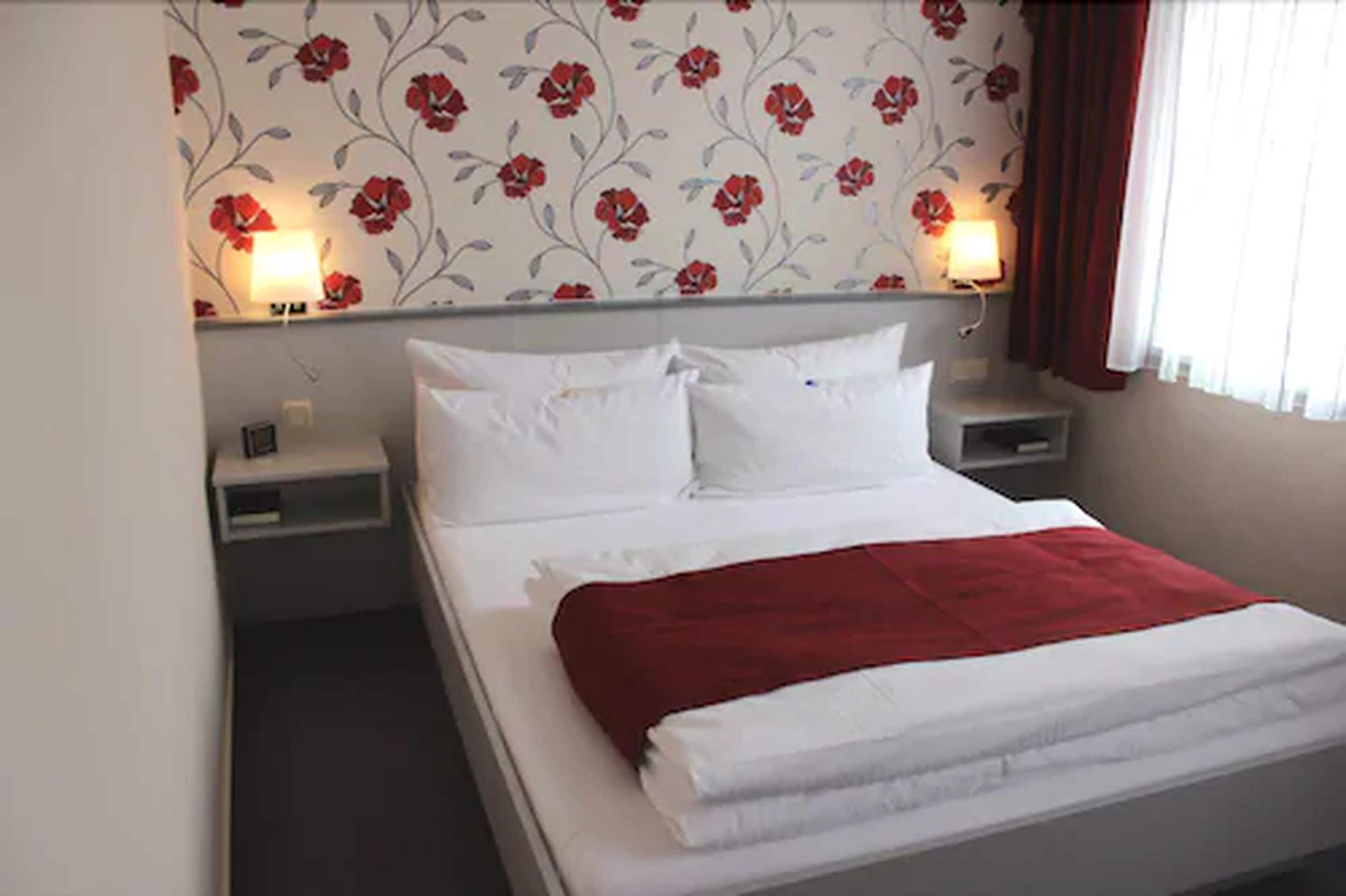 Una cama bien hecha con ropa de cama blanca y roja, dos mesitas de noche con lámparas y un estampado floral en la pared.