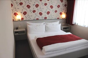 Una cama bien hecha con ropa de cama blanca y roja, dos mesitas de noche con lámparas y un estampado floral en la pared.
