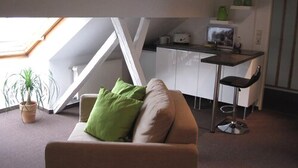 En kompakt opholdsstue med et tekøkken, en sofa med pude, et lille bord og en potteplante.