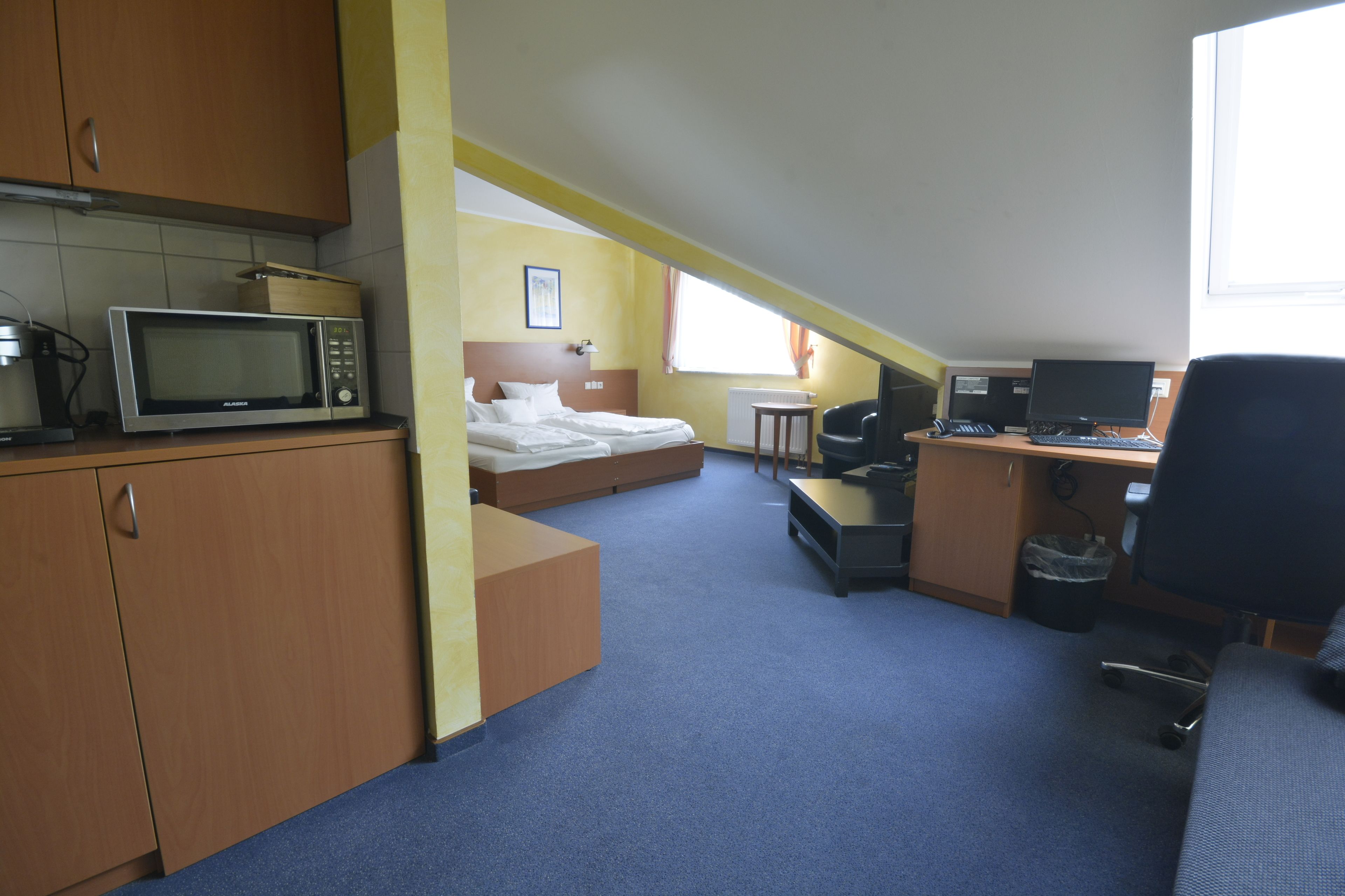 Ein kompaktes Hotelzimmer mit Küchenzeile, einem Bett, einem Schreibtisch mit Computer und einem Fenster für natürliches Licht.