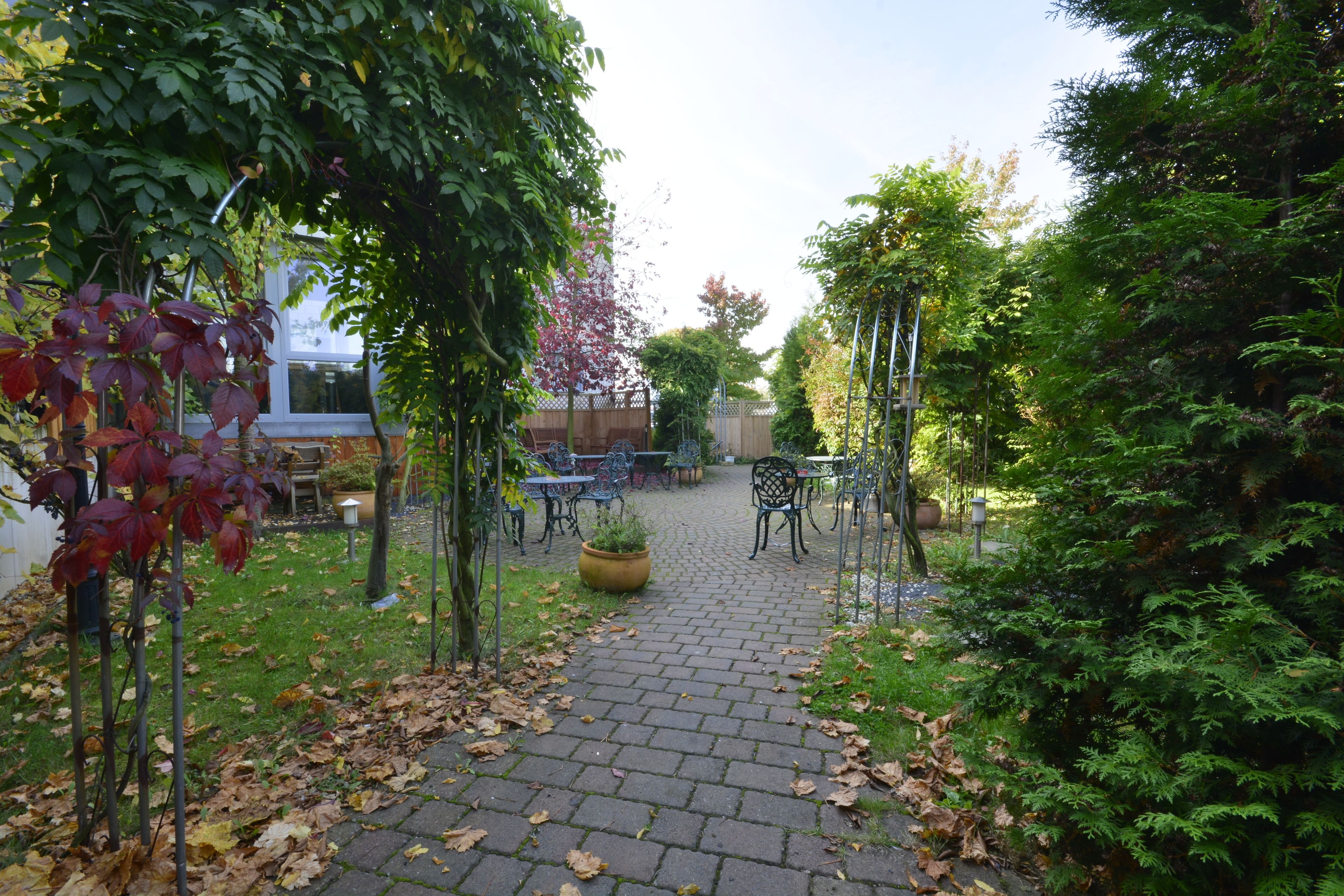 A paved garden path leading to outdoor seating with tables and chairs, surrounded by greenery and trees.