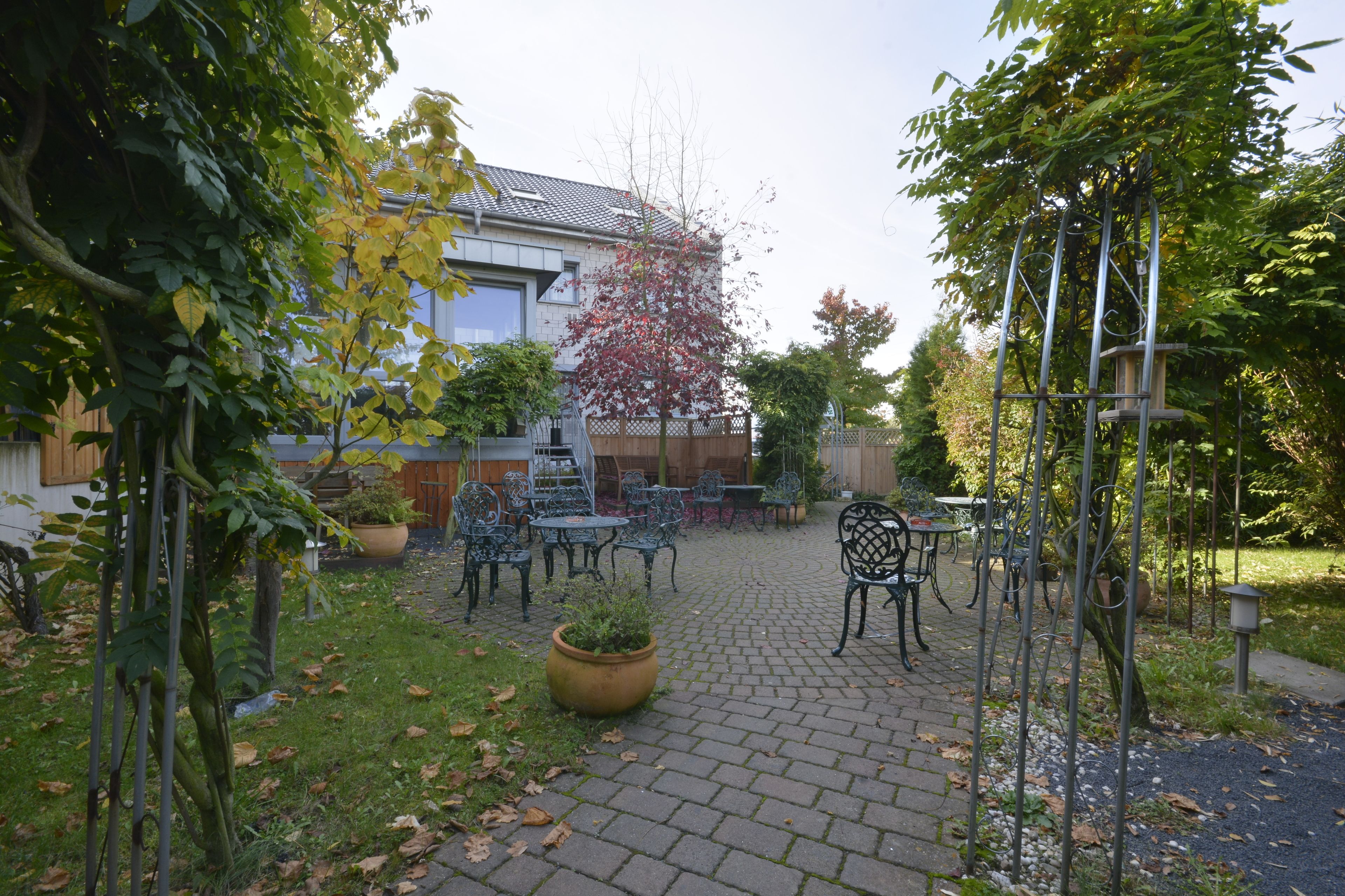 Ein Innenhof mit Pflasterweg, Metallstühlen und -tischen, umgeben von Grünpflanzen und einem Gebäude mit Ziegeldach.