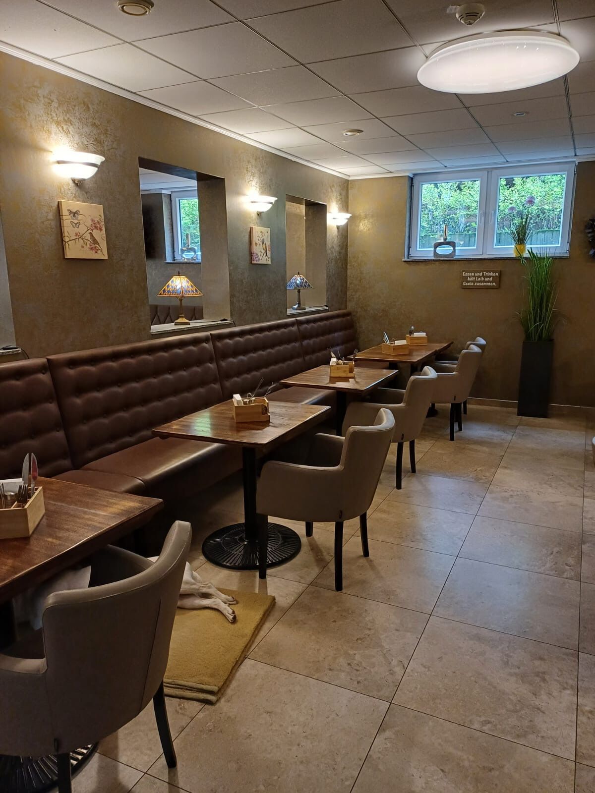 A restaurant with booths and tables, a tiled floor, and a window with a view of greenery.