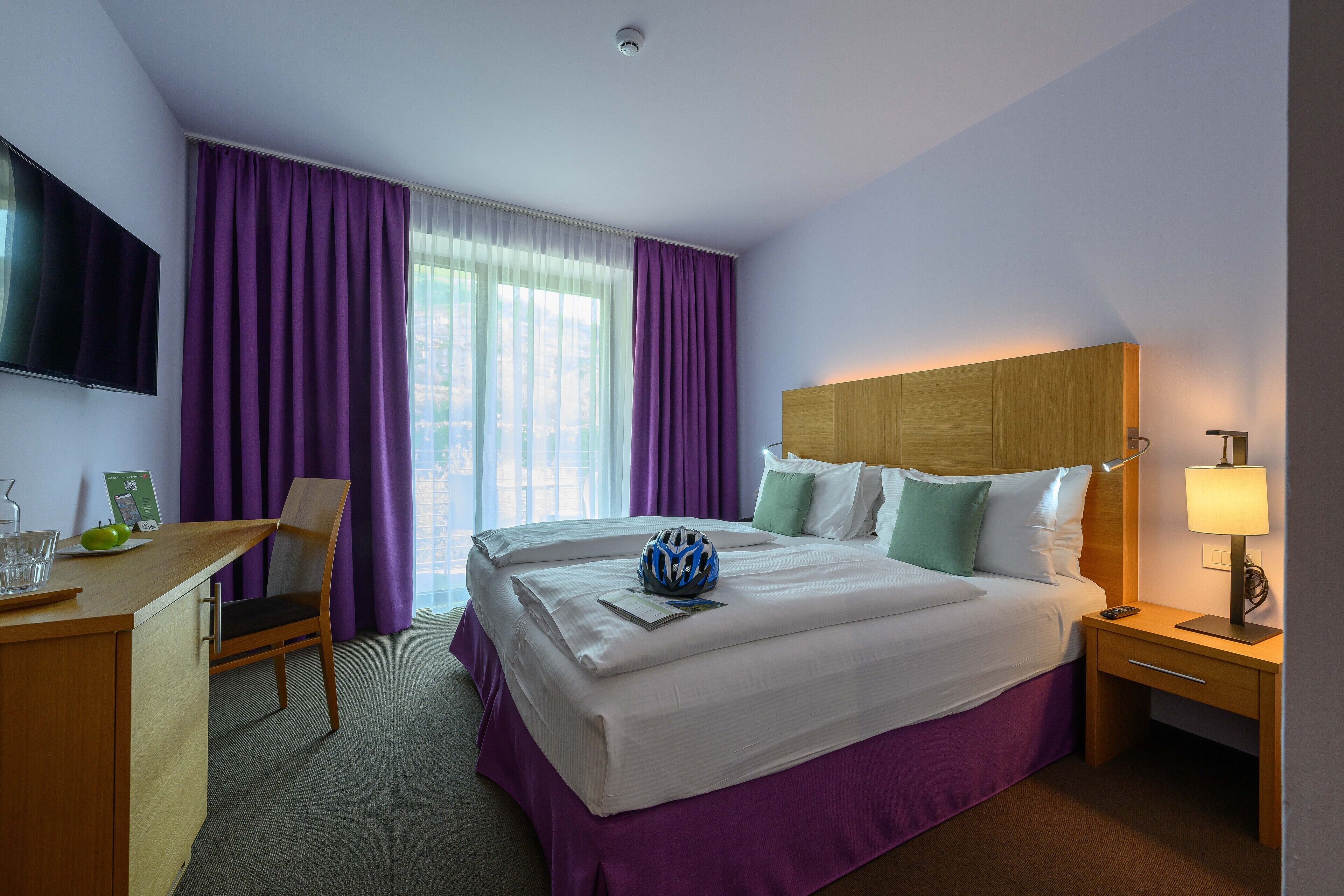 A hotel room with a bed, a desk, a chair, a TV, and a window with purple curtains.