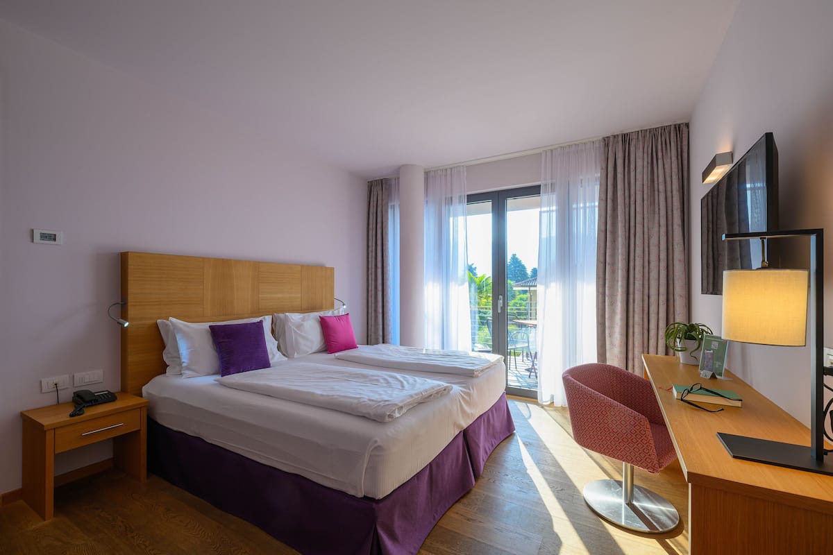 Een hotelkamer met een groot bed, een bureau met een lamp en uitzicht op de omgeving door een schuifdeur.