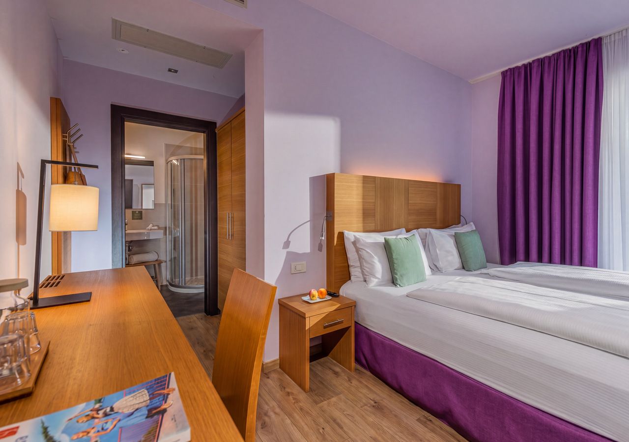 Una habitación de hotel con cama, mesita de noche, lámpara y vista al pasillo.