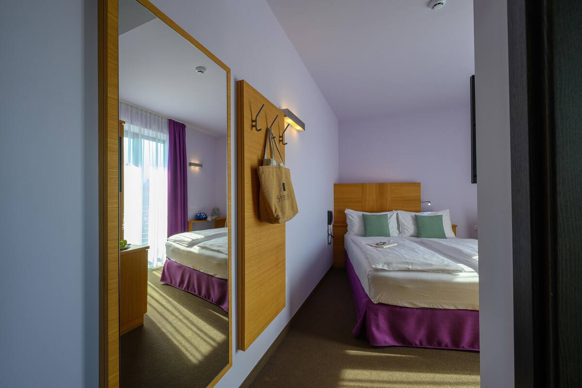 Een hotelkamer met een grote spiegel, een bed met een houten hoofdbord, een nachtkastje en een hangende tas.