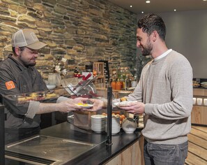 Two individuals at a buffet counter with plates of food, a stone wall background, and a variety of dishes and condiments.