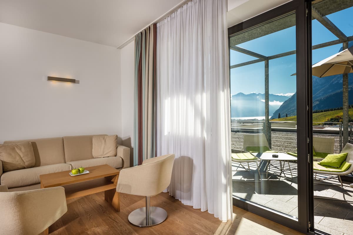 Een moderne woonkamer met een bank, salontafel en stoelen, grote ramen met gordijnen en uitzicht op bergen en een meer.