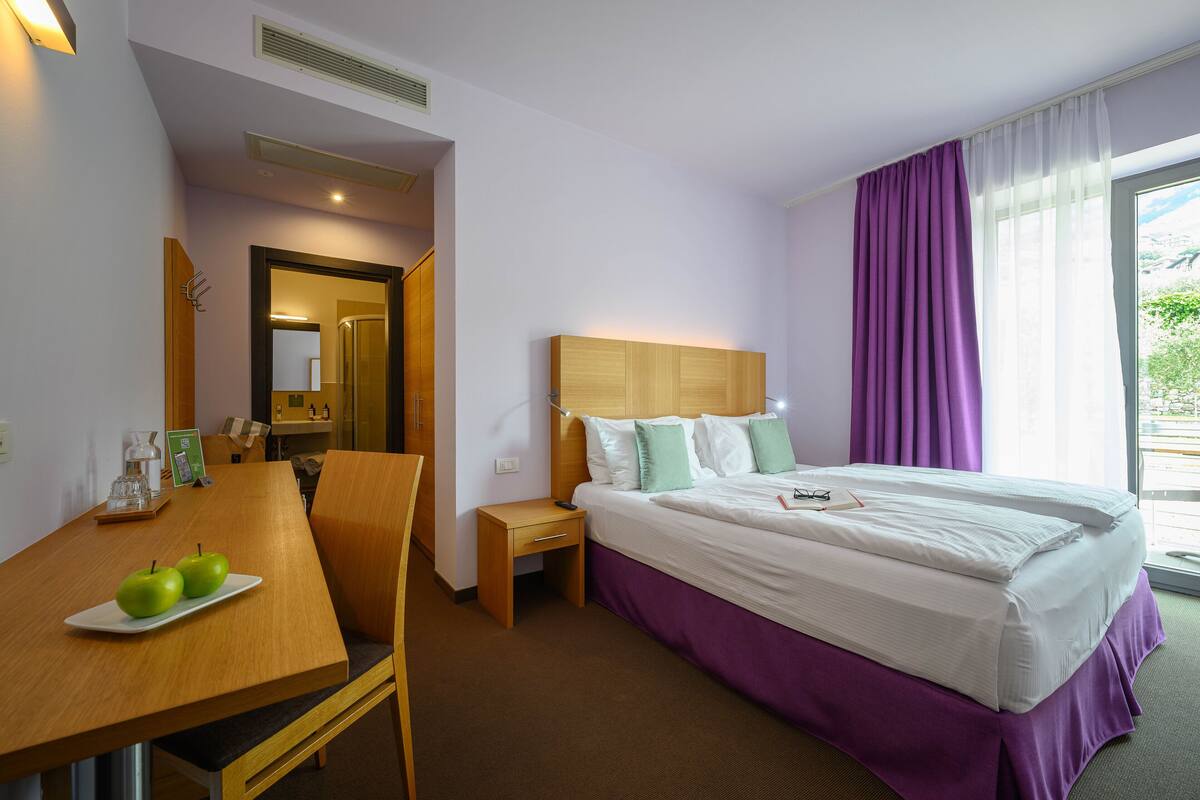 Een hotelkamer met een bed, een bureau met appels, een nachtkastje en een raam met gordijnen.