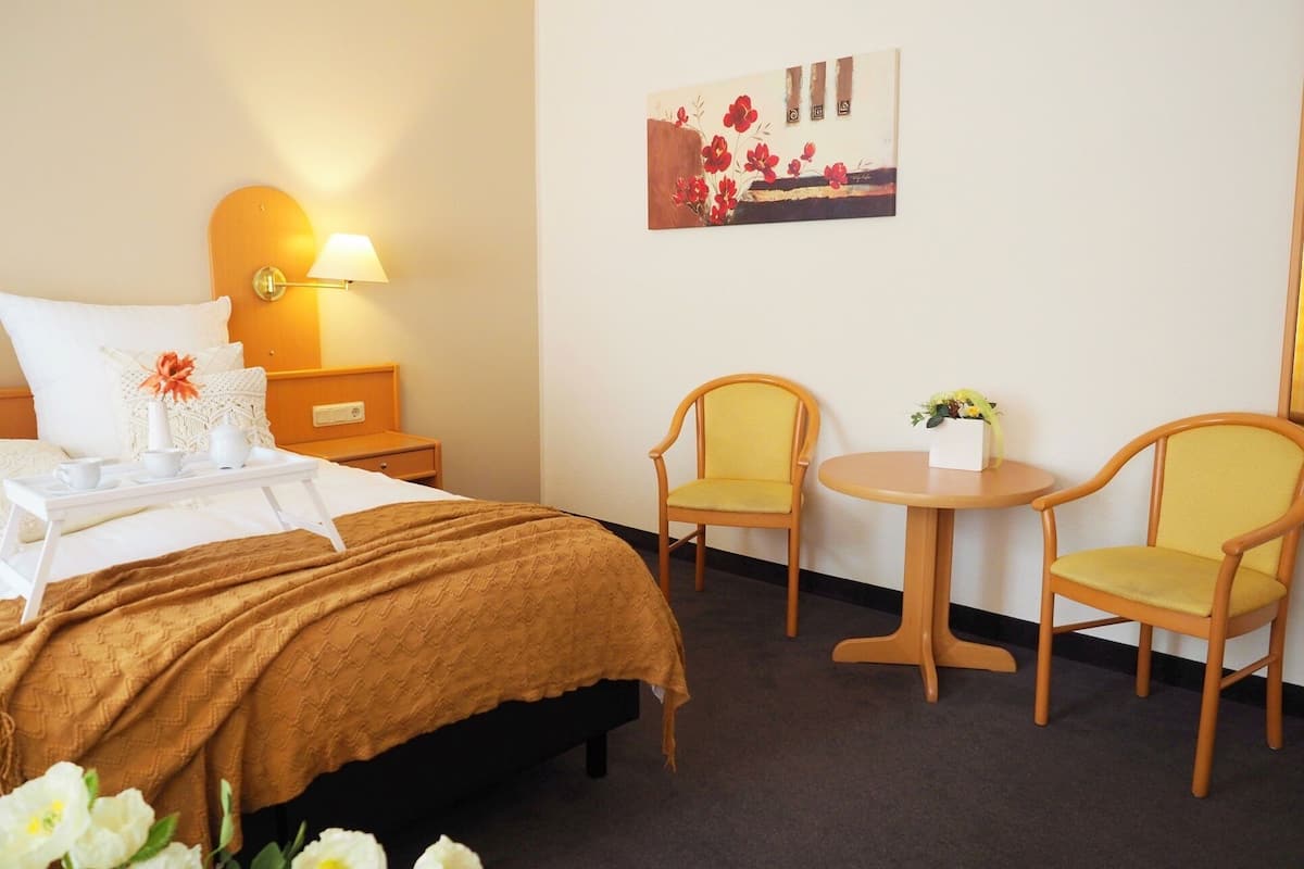 Une chambre d’hôtel comprenant un lit, une table de chevet, une lampe, deux chaises, une petite table et un tableau au mur.