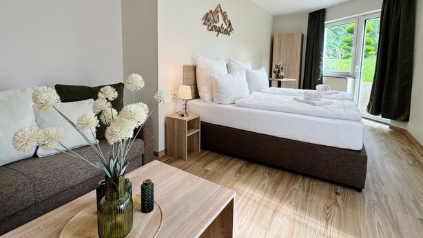 A hotel room with a bed, a sofa, a nightstand, a lamp, a vase with flowers, and a window with curtains. - Regiohotel Naturresort Ilsenburg  (Ilsenburg)