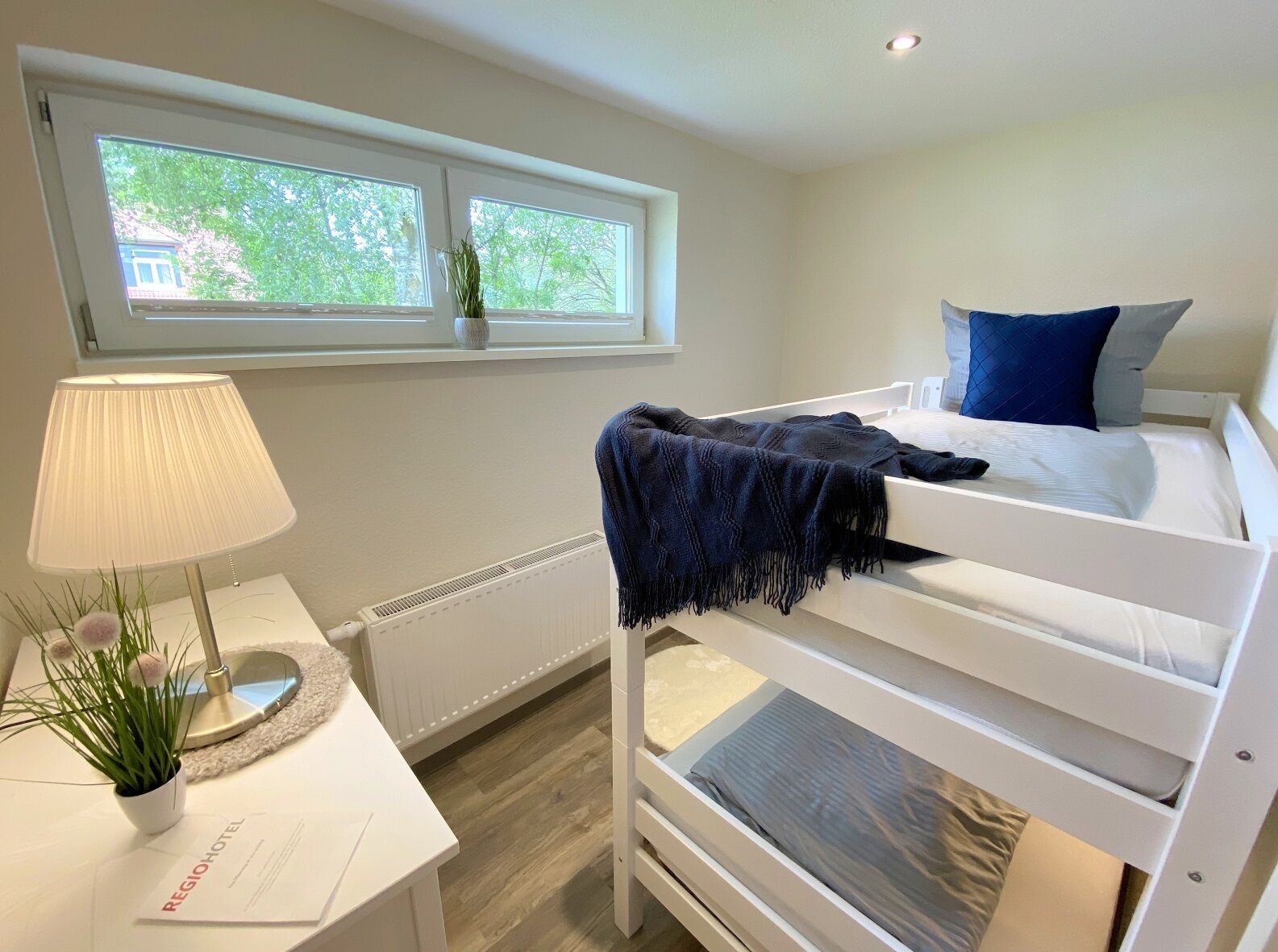 A bunk bed with a blue blanket, a bedside table with a lamp, and a window with a view of trees and a building.