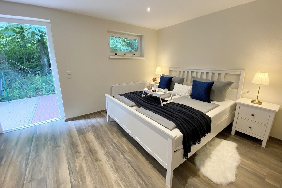 Une chambre moderne avec un lit blanc, deux tables de chevet, une petite fenêtre et une vue sur la verdure.