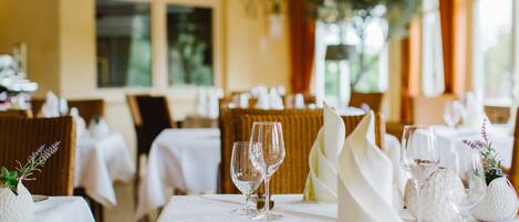 Comedor con mesas dispuestas para una comida, destacando manteles blancos, cubiertos y copas de vino.
