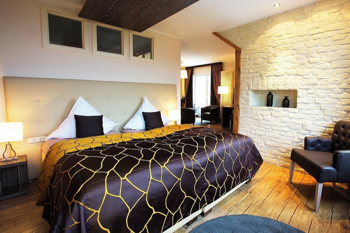 Une chambre d’hôtel avec un grand lit, un mur décoratif en pierre, un coin salon avec un fauteuil et une petite table.