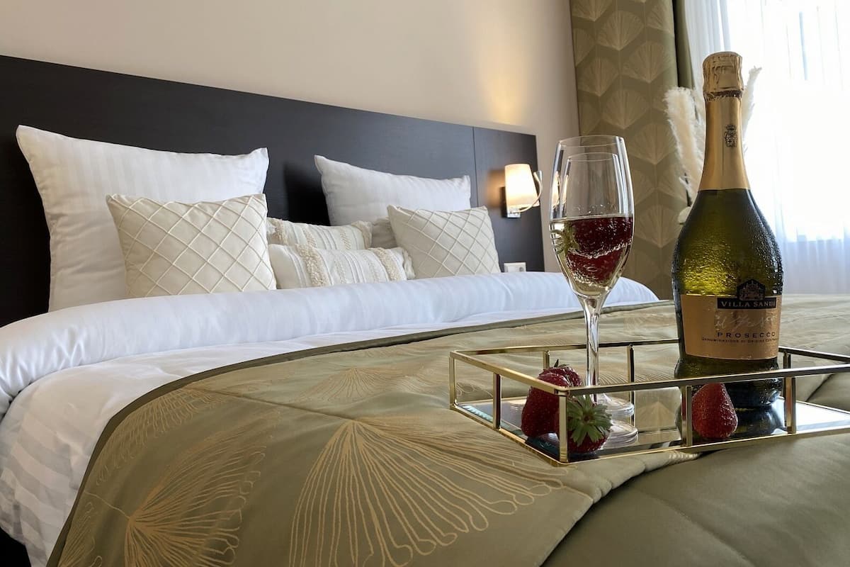 Un lit bien fait, avec du linge de lit blanc et des coussins décoratifs, une table de chevet sur laquelle se trouvent une bouteille de vin et un verre, et un plateau avec des fraises.