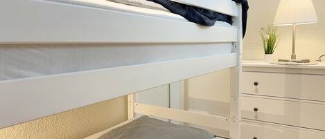 A bunk bed with a grey mattress and a white headboard. There is a bedside table with a lamp and a potted plant.