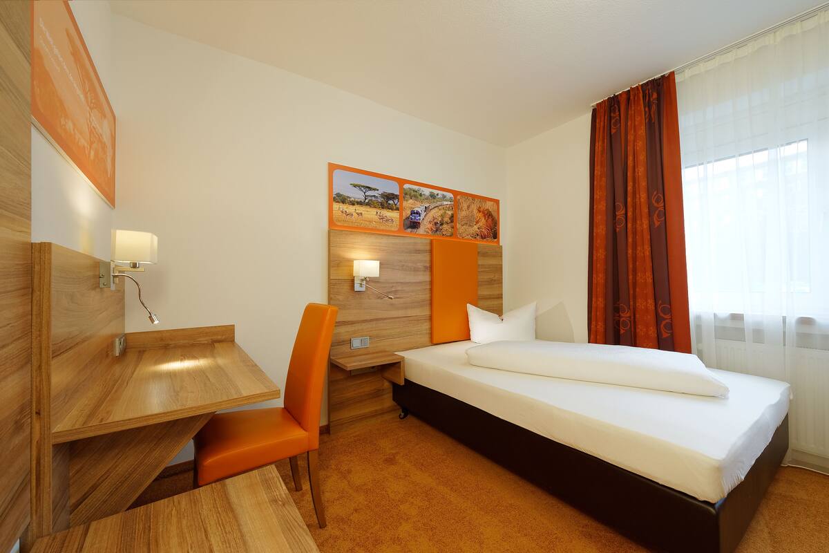 A hotel room with a wooden desk, orange chair, and a bed with white bedding.