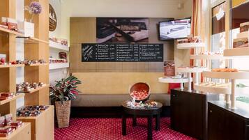 A cozy boutique hotel lobby with wooden shelves displaying chocolates and pastries, a comfortable sofa, a small table with a bowl of fruit, and a chalkboard menu listing food items.
