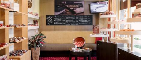 Eine gemütliche Hotelobby eines Boutique-Hotels mit Holzregalen, die Pralinen und Gebäck ausstellen, einem bequemen Sofa, einem kleinen Tisch mit Obstschale und einer Kreidetafel mit Speiseangeboten.