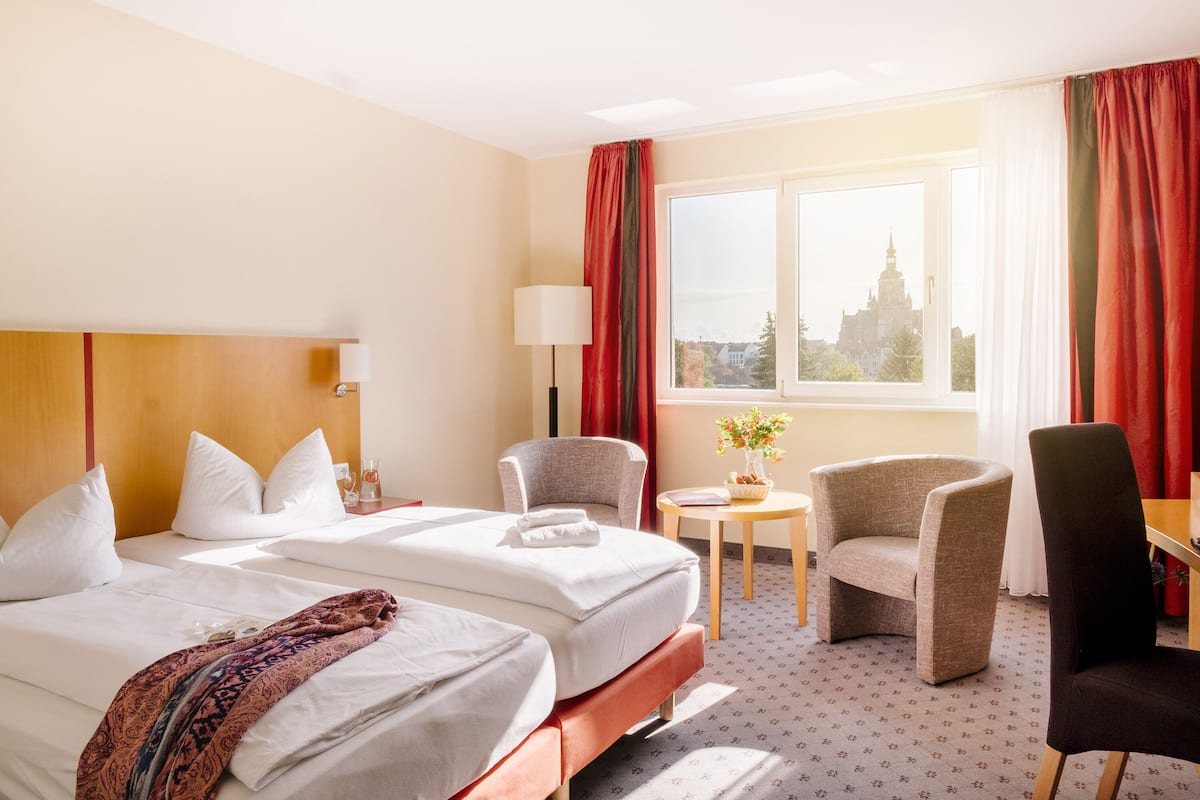 Habitación de hotel con dos camas, una mesita con un florero y vistas a la ciudad a través de un ventanal grande.