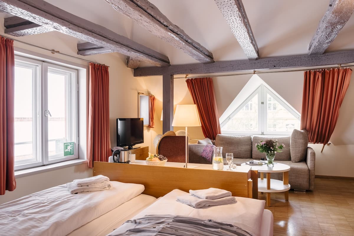 Habitación de hotel con cama, sofá, una pequeña mesa con un florero, lámpara y televisor.