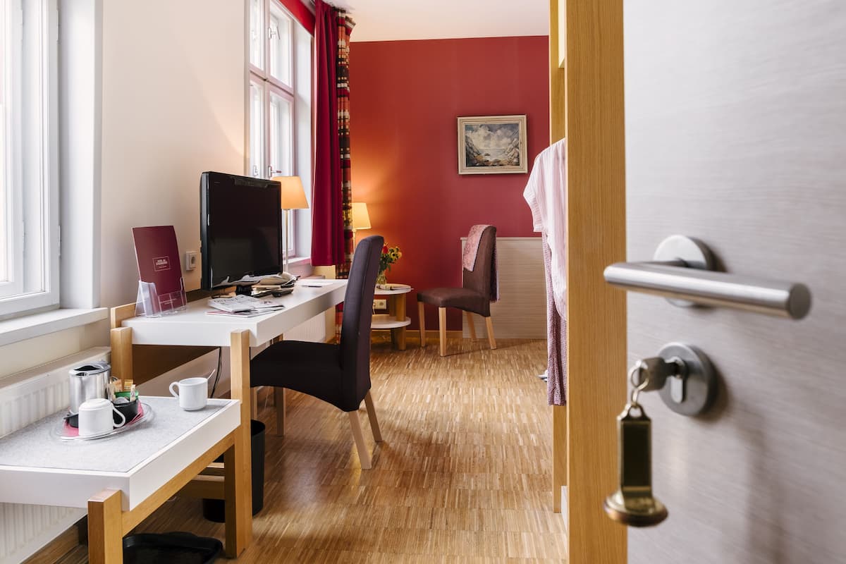 Habitación de hotel con suelo de madera, un escritorio con computadora, una silla, una pequeña mesa con lámpara y un cuadro en la pared.
